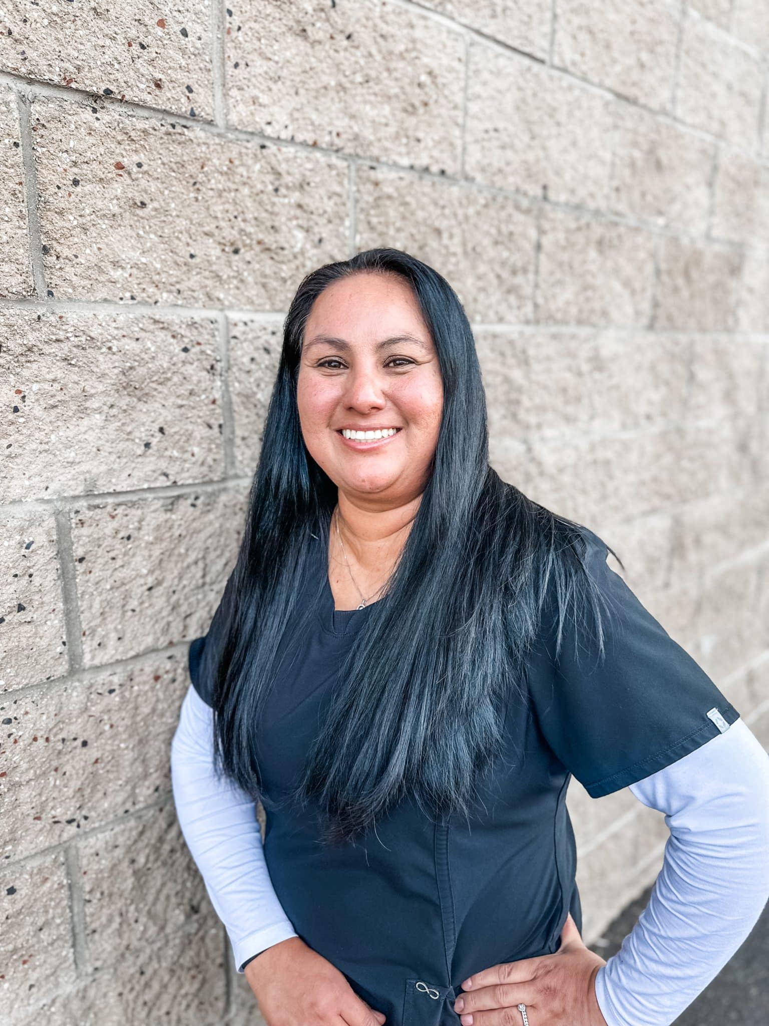Smiling person in scrubs standing against brick wall.